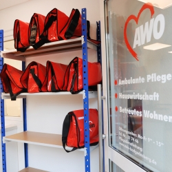 Eine offene Tür gibt den Blick auf mehrere rote Taschen des AWO-Pflegedienstes frei, die in einem Regal stehen.